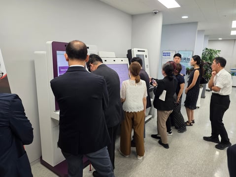 The wider group of delegates test out Dot Kiosk. 3 kiosks are in a row being demonstrated by Dot staff. 10 people in total are in this image - both make and female and all wearing business dress